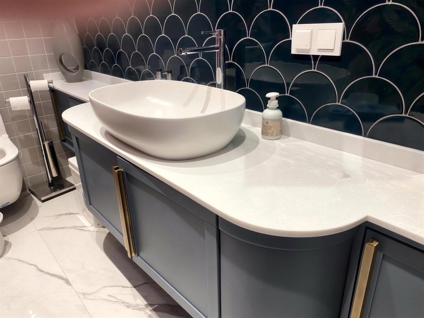 Gri-mavi lake kavisli gövdeli banyo dolabı, beyaz tezgâh, çanak lavabo ve pirinç kulplar